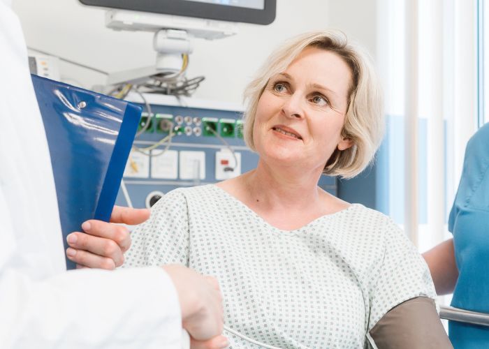 Mulher de meia-idade em uma consulta médica, usando avental hospitalar, conversando com um médico que segura uma prancheta, em ambiente de hospital.