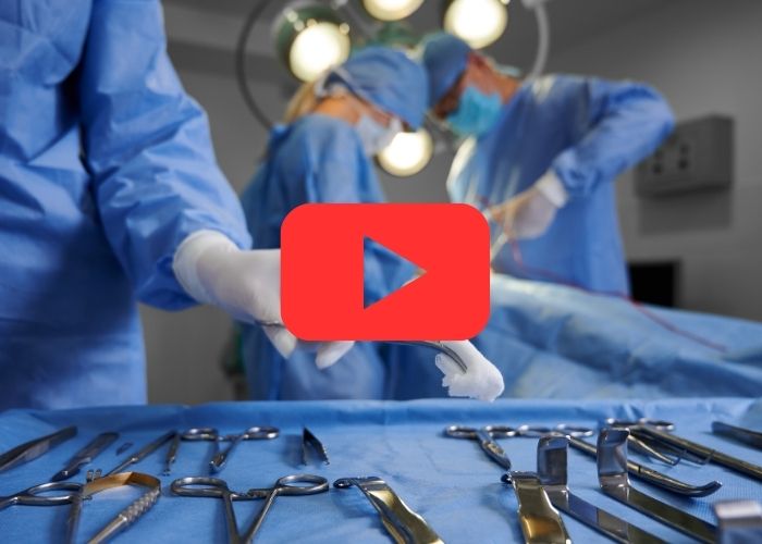 Cirurgiões vestidos de azul realizando uma cirurgia em ambiente esterilizado, com instrumentos cirúrgicos dispostos sobre uma mesa ao fundo e ícone do YouTube sobreposto.