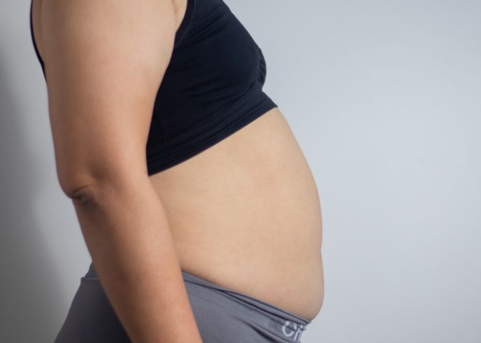 Mulher com sobrepeso em perfil, vestindo top preto e calça cinza, destacando a região abdominal saliente.