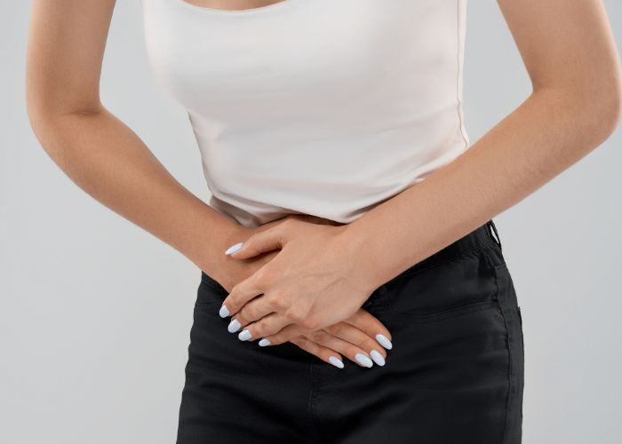 Mulher com expressão de dor, segurando a parte inferior do abdômen com as duas mãos, vestindo blusa branca e calça preta.