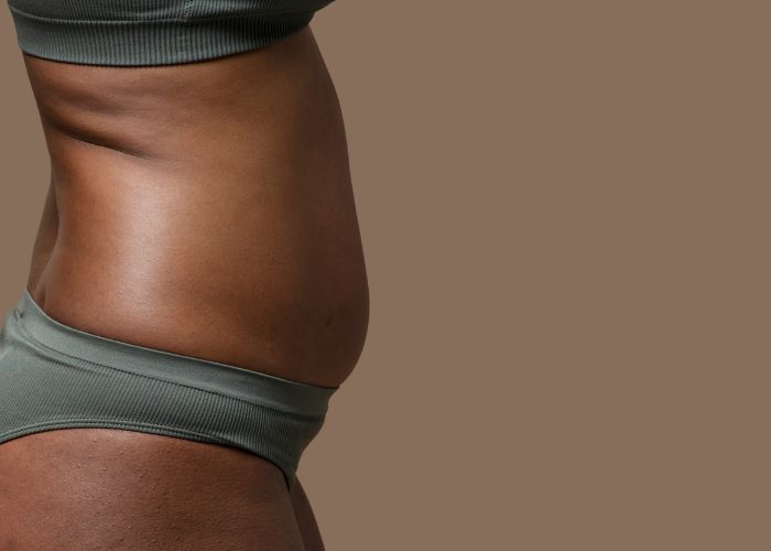 Close do abdômen de uma mulher em roupa de treino, evidenciando leve protuberância abdominal, com fundo marrom neutro.