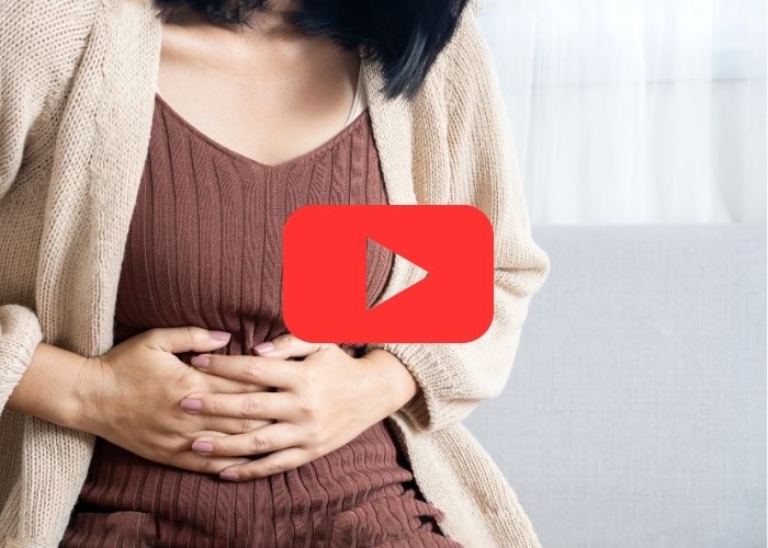 Mulher sentada com as mãos sobre o abdômen, indicando dor ou desconforto abdominal, com ícone do YouTube sobreposto.