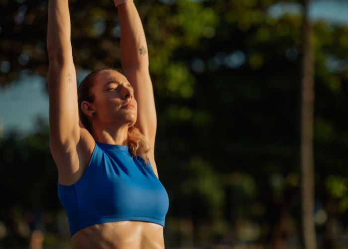 Mulher com roupa esportiva azul faz alongamento ao ar livre, com olhos fechados e expressão relaxada, em ambiente ensolarado e arborizado.