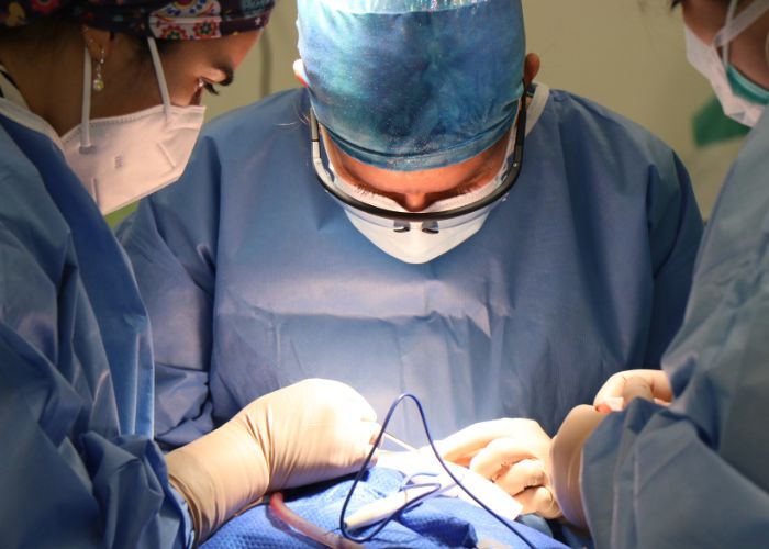 Pessoa vestida com roupa cirúrgica e máscara realiza uma cirurgia, acompanhada por outros profissionais de saúde na sala de operação.