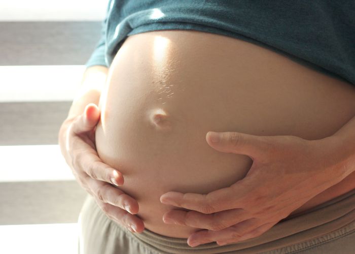 Mulher grávida segura a barriga com as duas mãos, destacando o umbigo projetado, sugerindo uma possível hérnia umbilical pós-gestação.