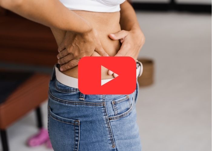 Mulher com as mãos na região lombar, aparentando dor; veste jeans e top branco em ambiente interno.