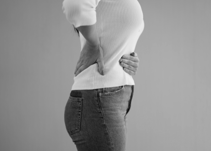 Mulher com as mãos na região lombar e abdominal, aparentando desconforto ou dor, vestindo blusa branca e calça jeans em fundo neutro.