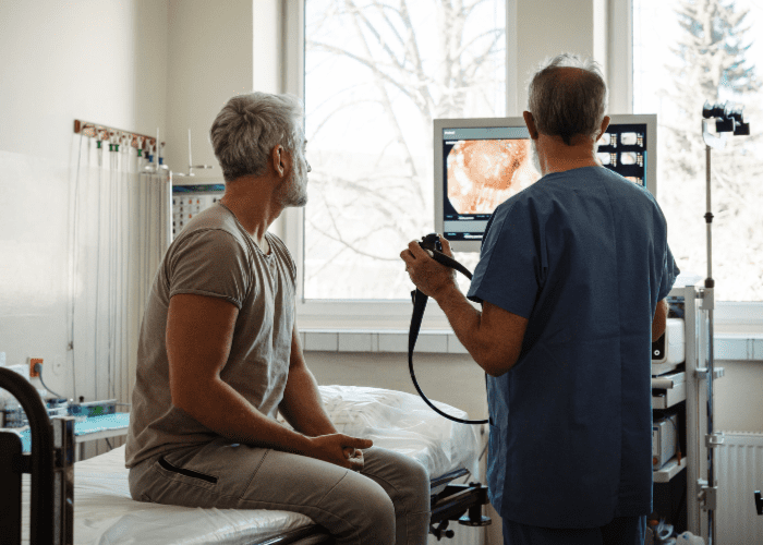 Médico mostra imagens de colonoscopia em um monitor enquanto paciente sentado na maca observa, em consultório iluminado por janelas amplas.