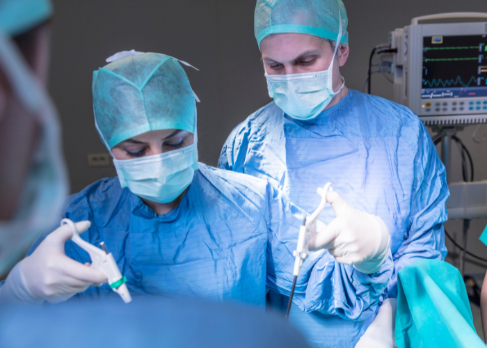 Equipe médica realiza cirurgia laparoscópica em centro cirúrgico, utilizando instrumentos e monitoramento hospitalar ao fundo.