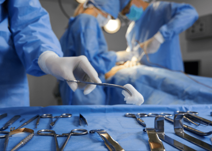 Equipe cirúrgica em procedimento hospitalar, com instrumentos sobre mesa estéril e profissional segurando gaze durante a cirurgia.