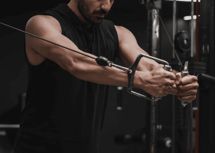 Homem realizando exercício de força em máquina de cabos na academia, com foco nos braços e esforço muscular durante o movimento.