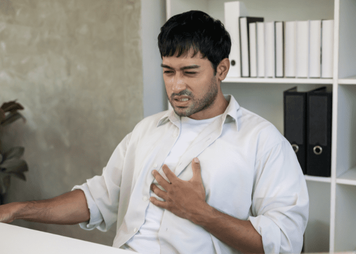 Homem sentado em ambiente interno, com a mão no peito e expressão de desconforto, sugerindo azia ou dor torácica associada ao refluxo.