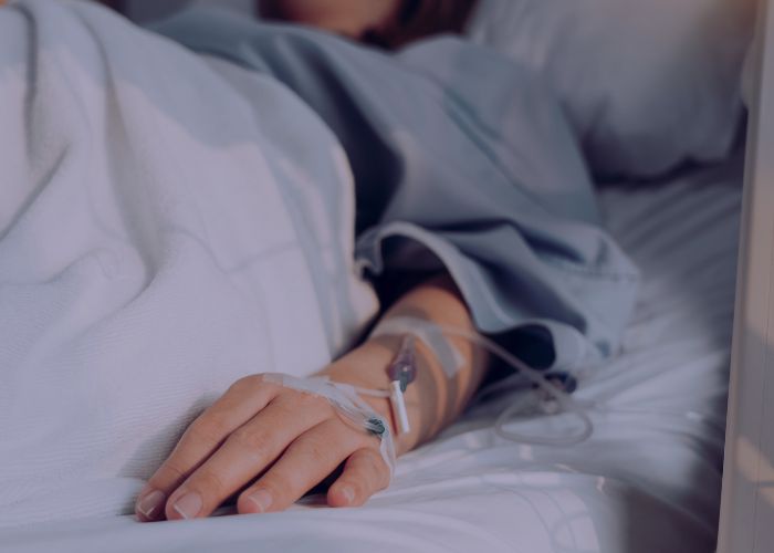 Paciente deitado em leito hospitalar com acesso venoso na mão, em ambiente de recuperação e cuidados médicos.