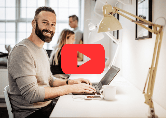 Homem sorridente trabalhando em laptop em escritório, sentado à mesa com colegas ao fundo em ambiente profissional iluminado.