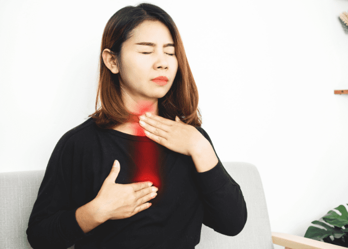 Mulher sentada com expressão de desconforto, levando a mão ao pescoço e ao peito, indicando sensação de dor ou queimação.