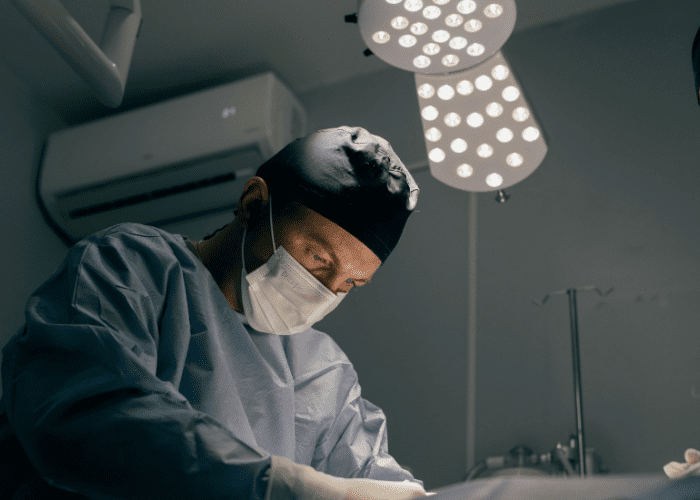 Cirurgião em sala operatória realiza procedimento sob iluminação cirúrgica, usando máscara e vestimenta estéril.