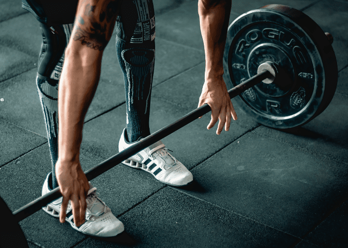 Pessoa segurando barra com pesos durante treino de musculação em academia, com foco nas mãos, pernas e anilhas.