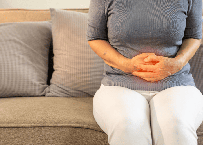Pessoa sentada em um sofá com as mãos sobre o abdômen, demonstrando desconforto abdominal, com destaque em vermelho na região da barriga.